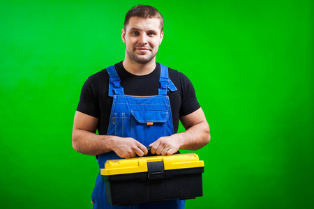 一个强壮的木匠穿着黑色T恤和蓝色建筑连衣裙，微笑着拿着一个盒子，肩膀上有建筑工具，背景是绿色的孤立的。