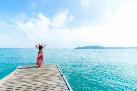 夏日。 微笑的女人放松，穿着红色的衣服时尚站在海上蓝天背景上的木桥上。 旅行和度假。 自由概念