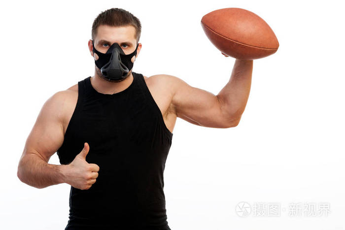 一个年轻的运动员穿着运动衫训练黑色面具，手里拿着一个橄榄球球，手里拿着拇指在白色的孤立背景上