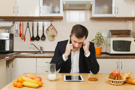 疲惫的年轻商人穿着西装, 衬衫, 吃早饭, 坐在桌子上, 用平板电脑, 在手机上聊天, 在光厨房吃水果。健康的生活方式。在家做饭