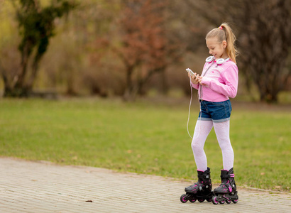 溜冰鞋和小女孩美丽的微笑小女孩在线滑冰通过公园和使用智能手机.