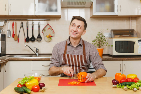 英俊的微笑高加索年轻人在一个围裙, 棕色衬衫坐在餐桌上, 削减蔬菜沙拉用刀在光厨房。节食概念。健康的生活方式。在家做饭。准备食物