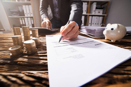 s Hand Signing Document With Stacked Golden Coins On Wooden Desk