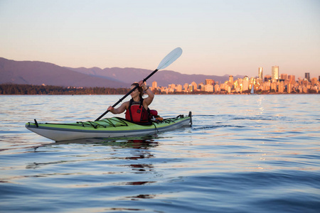 美丽的女人海上皮划艇在海洋中，在五彩缤纷和充满活力的日落与城市天际线在后面。 摄于杰里科温哥华不列颠哥伦比亚省加拿大。