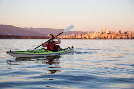 美丽的女人海上皮划艇在海洋中，在五彩缤纷和充满活力的日落与城市天际线在后面。 摄于杰里科温哥华不列颠哥伦比亚省加拿大。