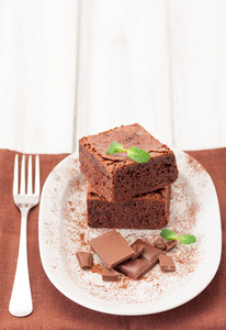 巧克力布朗尼方形块在白色的盘子装饰与薄荷叶和可可粉白色复古木背景。美国传统的美味甜点。关闭摄影