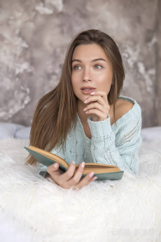 年轻美丽的高加索女人坐在床上看书. 她穿着休闲装:蓝色毛衣和牛仔裤.