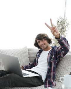 快乐的人，与笔记本电脑坐在沙发上，显示他的手一个胜利的手势