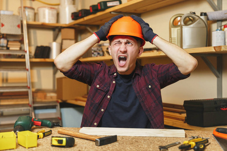 疯狂进取愤怒的高加索年轻人在格子衬衫, 黑色 t恤, 橙色防护头盔, 在木工车间的手套工作在木制桌子地方用木锤不同的工具