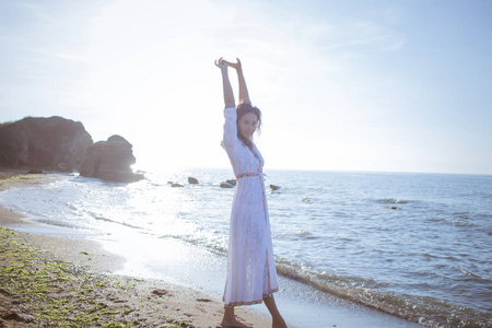 年轻的黑发女人穿着漂亮的白色连衣裙在早晨的夏天海滩上摆姿势