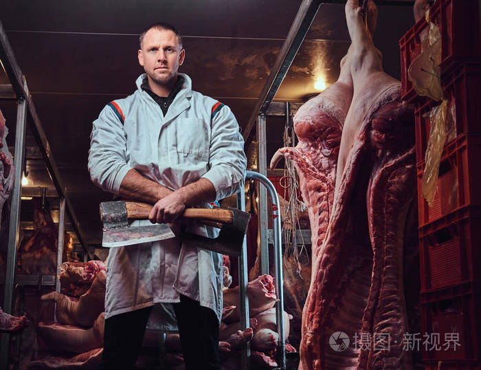 一个穿白色厨房长袍的屠夫的肖像在一家肉厂里拿着一把斧头