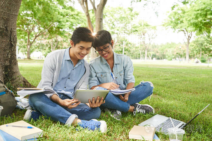 快乐的大学生坐在公共公园里在绿色草坪上共同编写项目教科书和教科书