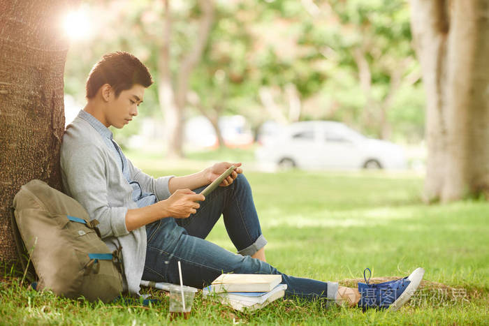 自信的亚洲学生坐在树下,用数字平板电脑做学校项目