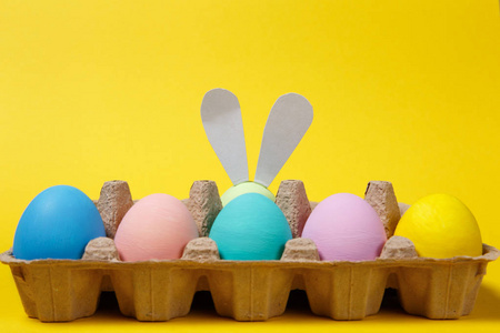 一排五颜六色的蜡笔声道画复活节彩蛋与有趣的兔子耳朵在纸板托盘上的黄色背景卡。复活节快乐的概念。广告的复制空间。与地方为文本