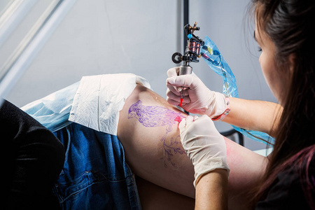 关闭主纹身绘制红色油漆在客户纹身。 纹身艺术家拿着一台无菌手套的金属纹身机，在专业的蓝色垫子上工作。