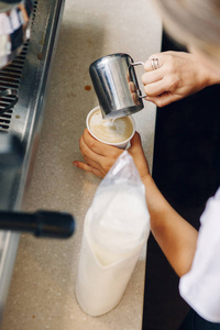 高加索咖啡师手工制作卡布奇诺的特写镜头。 服务员或小企业主在咖啡纸杯里倒热牛奶。