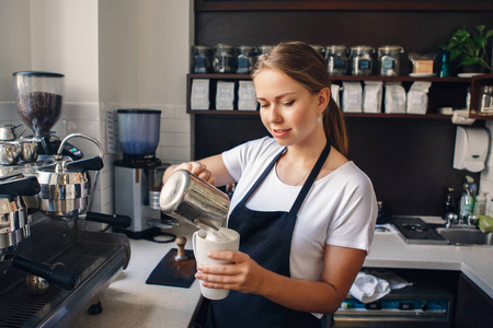 微笑的年轻白种人女咖啡师在围裙倒牛奶咖啡的肖像。 企业主的概念。