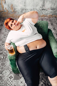 超重妇女坐在扶手椅上看电视，喝啤酒，肥胖和不健康的生活方式观念