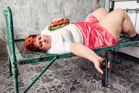 胖女人躺在生锈的床上吃三明治，看电视，不健康的生活方式和肥胖观念