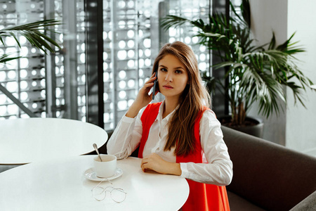 严格的商务女士坐在咖啡馆的一张小桌子上，在智能手机上交谈，喝咖啡。 在商务中心午餐时穿着白色衬衫的女人。
