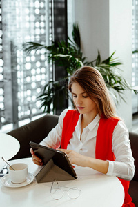 严格的商务女士坐在咖啡馆的一张小桌子上，在平板电脑上使用互联网，喝咖啡。 在商务中心午餐时穿着白色衬衫和红色夹克的女人。