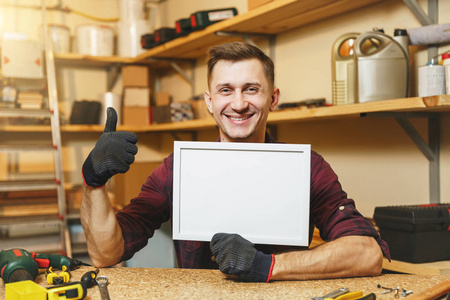 英俊的高加索青年男子在格子衬衫, 在木工车间的手套工作在木制表的地方与空白框架, 不同的工具。空的地方为文本。广告复制空间
