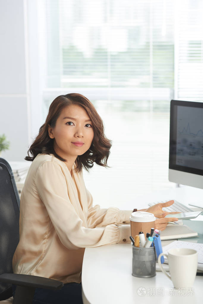 亚洲女商人坐在办公桌前,手里拿着平板电脑,看着相机照片