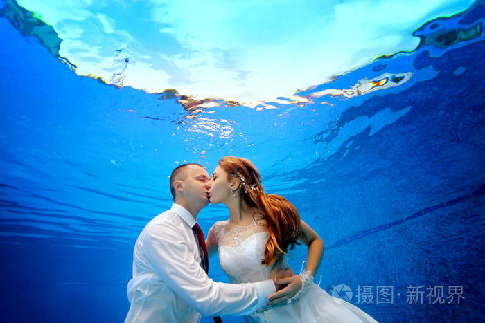 新娘和新郎在婚礼礼服拥抱和亲吻水下在水池在蓝色背景。关闭。景观定位。在游泳池水下射击