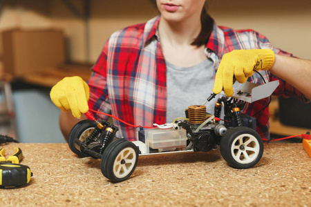 年轻女子在格子衬衫, 灰色 t恤, 黄色手套制作玩具车铁模型建设者, 在车间工作在木桌地方用不同的工具。电工用万用表
