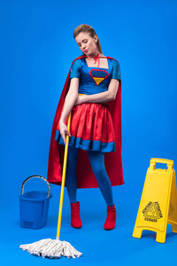 穿着超级英雄服装的迷人女人，小心标志桶和拖把，用蓝色隔离清洁
