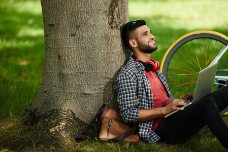 微笑的年轻人坐在树下，膝盖上放着笔记本电脑，抬头看着