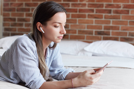 侧面肖像严肃的女性听音乐或新闻在电台网站上使用现代无线耳机添加多媒体文件，检查邮件姿势在她的晨床上穿着条纹衬衫。