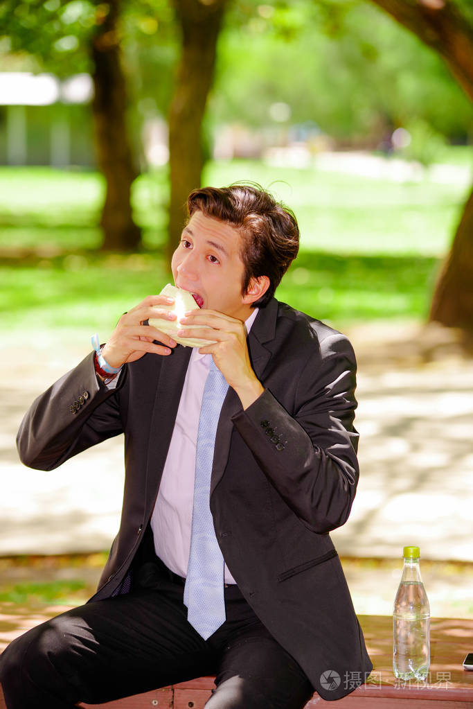 英俊年轻的生意人穿着西装, 在户外吃三明治, 在一个模糊的公园背景