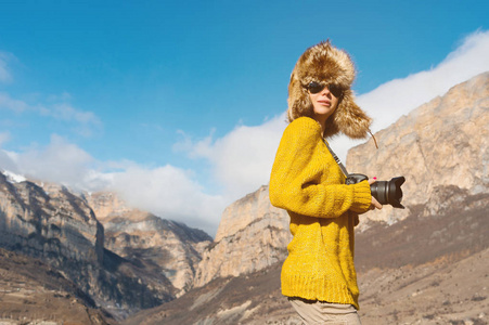 一个女孩摄影师在太阳镜和一个大的毛皮帽子和黄色针织毛衣站在与高岩石的背景, 在峡谷与单反相机她在手