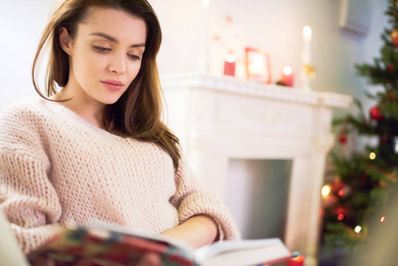 一位身穿毛衣坐在舒适的沙发上沉浸在阅读有趣的书客厅内部装饰背景圣诞庆祝活动中的迷人女人的画像