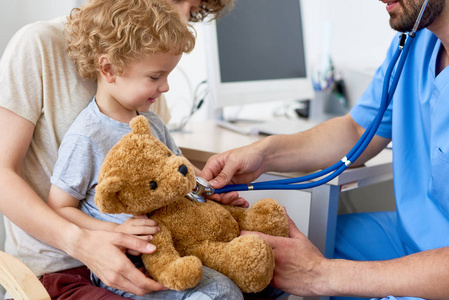 可爱的卷发孩子坐在医生办公室的母亲腿上，拿着泰迪熊玩具，儿科医生用听诊器听心跳