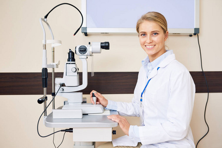 金发女眼科医生坐在裂隙灯机旁,摆姿势看镜头,开心地笑着照片