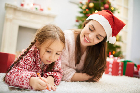 可爱的小女孩裹着写给圣诞老人的信，躺在壁炉旁的地毯上，她迷人的母亲带着迷人的微笑，盯着她