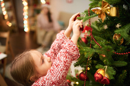 深色头发的小女孩的肖像，穿着针织毛衣，站在脚趾上装饰圣诞树，背景模糊