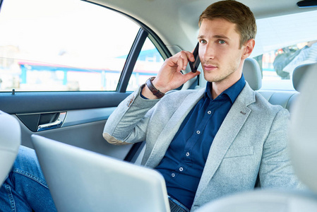 成功的年轻商人通过电话和笔记本电脑坐在豪华汽车的后座上的肖像。