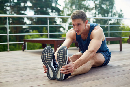 自信的年轻运动员坐在宽敞露台的木地板上，在密集锻炼前做伸展运动，背景风景如画