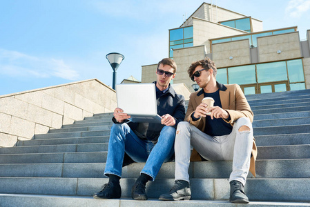 两位英俊的年轻人在户外使用笔记本电脑，坐在现代玻璃建筑的台阶上，享受着咖啡