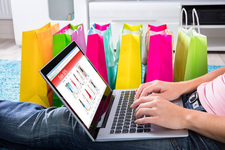 女人坐在地毯上，用笔记本电脑上网购物，还有五颜六色的购物袋