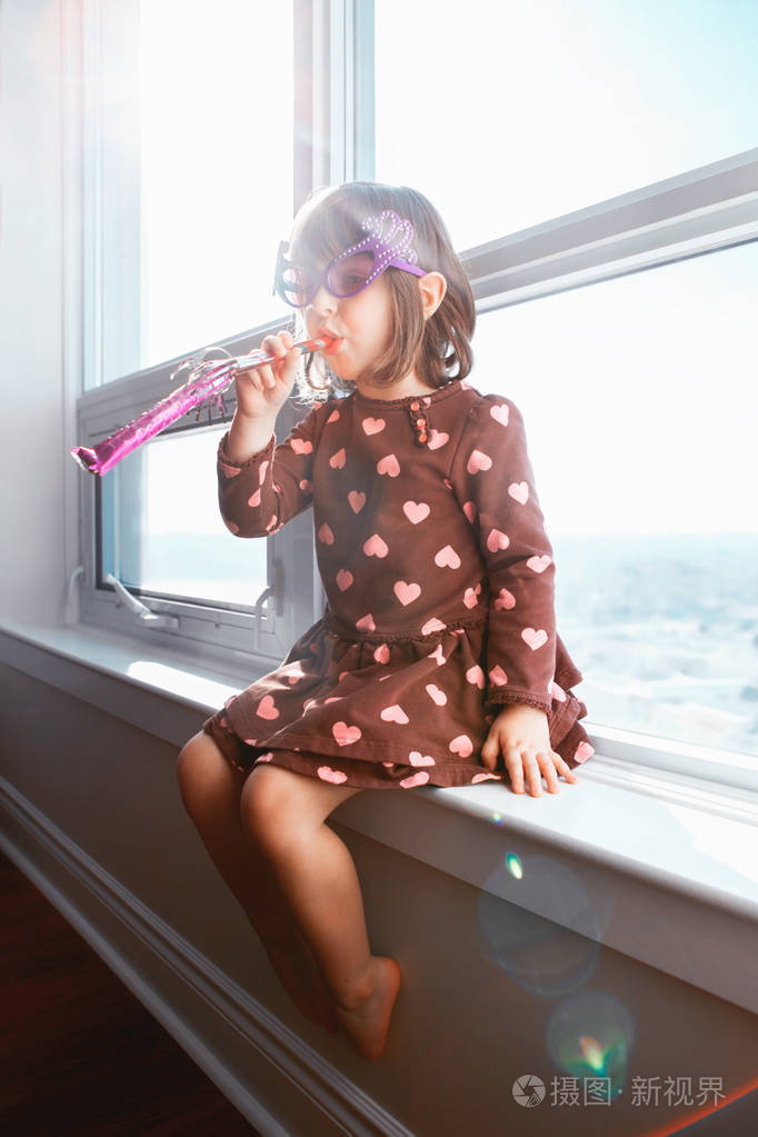 白种人女孩的肖像坐在窗台上在家里吹哨子庆祝生日.