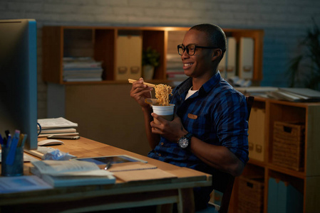 快乐的年轻商人在电脑上看电影,晚上独自坐在开放式办公室里吃中国菜