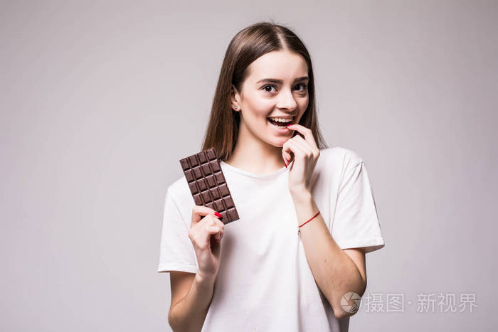 一个美丽的年轻女人咬着一块乳白色的巧克力，嬉戏，看着镜头，然后在灰色中孤立地吃