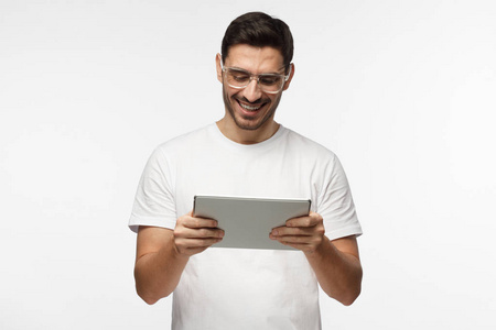 工作室特写照片上的年轻男子在灰色背景上的隔离戴着透明的眼镜, 聚精会神地看着平板电脑的屏幕他抱着, 高兴地笑着, 笑
