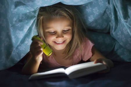 甜美美丽的金发碧眼的小女孩6到8岁以下的床上盖看书在黑暗中, 晚上与火炬轻微笑快乐