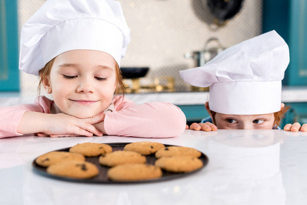 可爱的快乐孩子戴着厨师帽，看着桌上美味的饼干