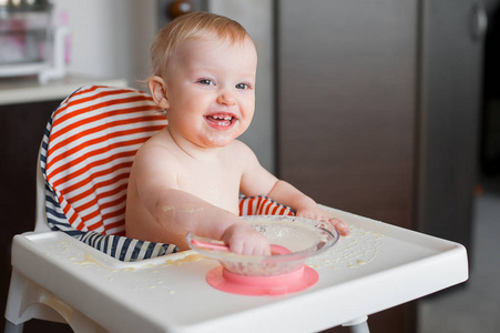 小可爱的女孩坐在高高的椅子和学习用勺子自己吃。逗人发笑的愉快的婴孩弄脏食物在桌